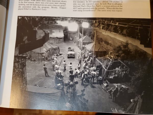 outdoor shot looking down on a soundstage made up like the MASH camp, with painted scenery backdrops and a number of actors on set