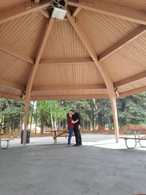A man and woman in ordinary street clothes stand beneath a high wooden pavilion, kissing. He is much taller and she is standing on her tiptoes. Trees and summer background.